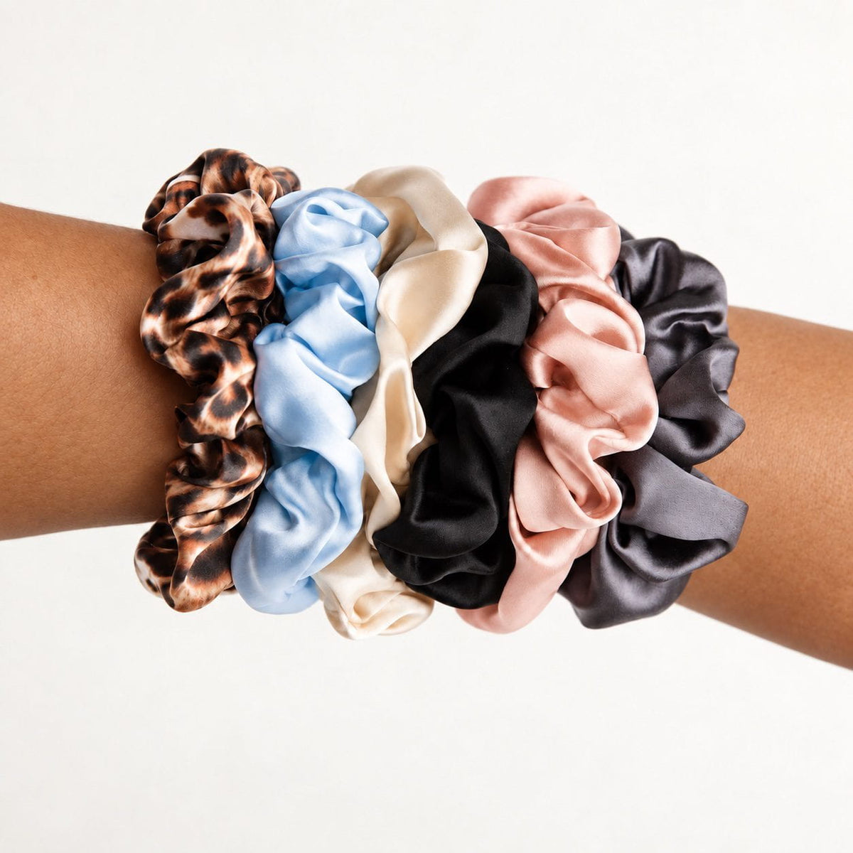 Six NIGHT Classic silk scrunchies worn on an arm in Leopard, Sky Blue, Champagne, Black, Blush, and Gunmetal