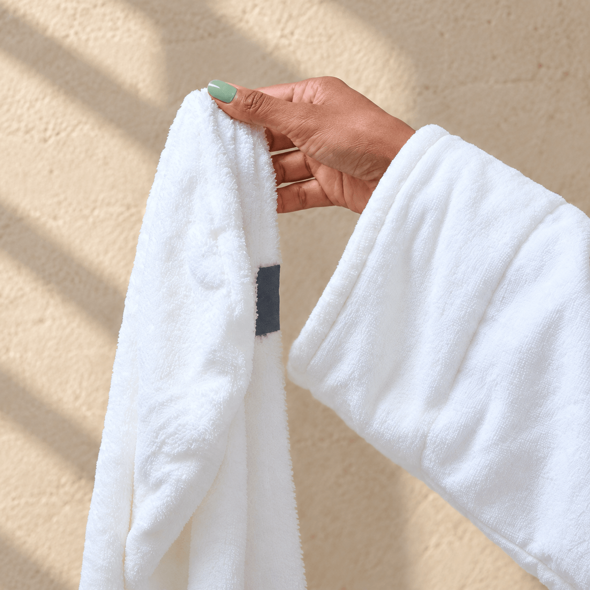 Close-up of a hand with light sage-green nails holding the white NIGHT microfiber hair towel against a warm beige background.