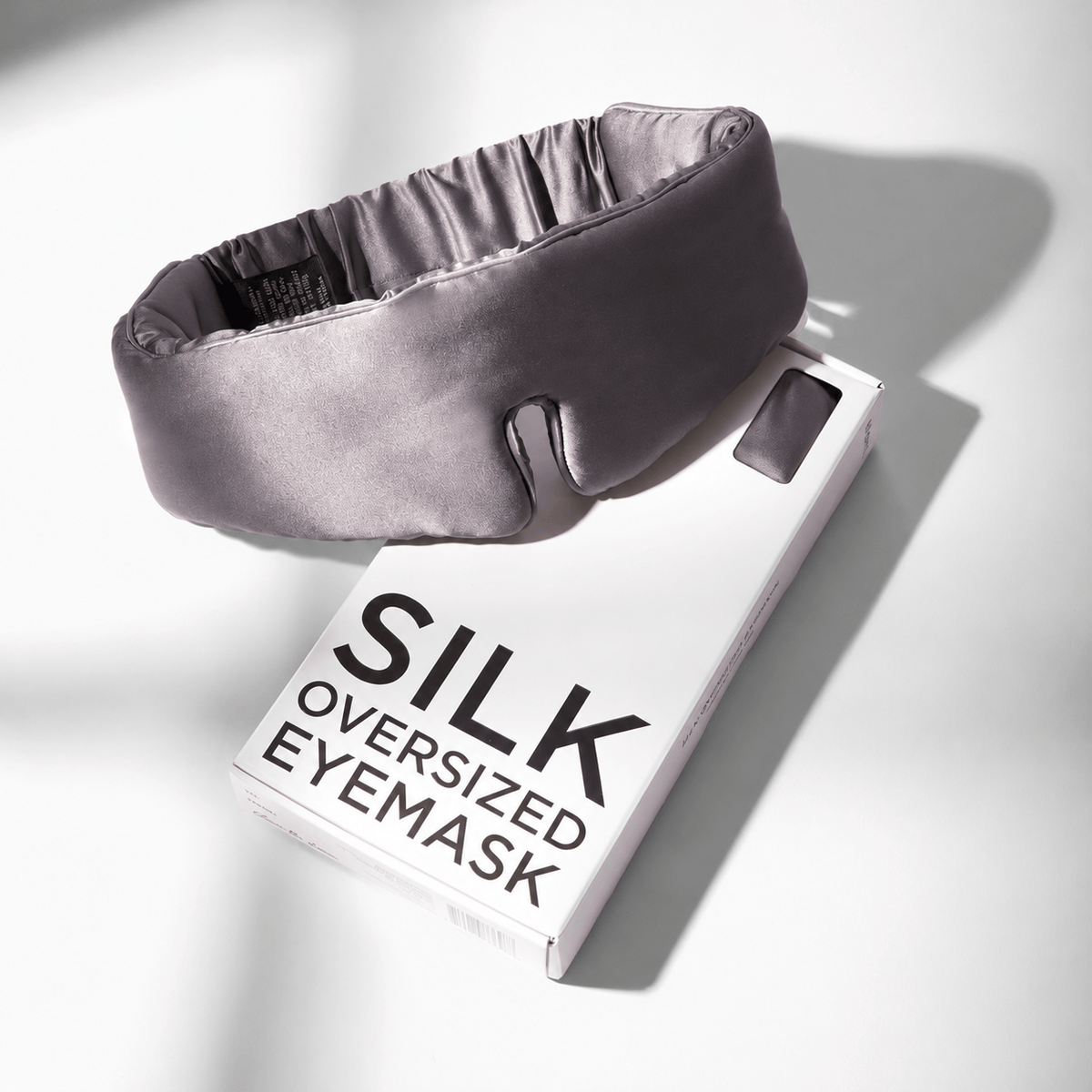 Gunmetal oversized silk sleep mask resting on its white SILK OVERSIZED EYEMASK gift box, showing the gathered adjustable band