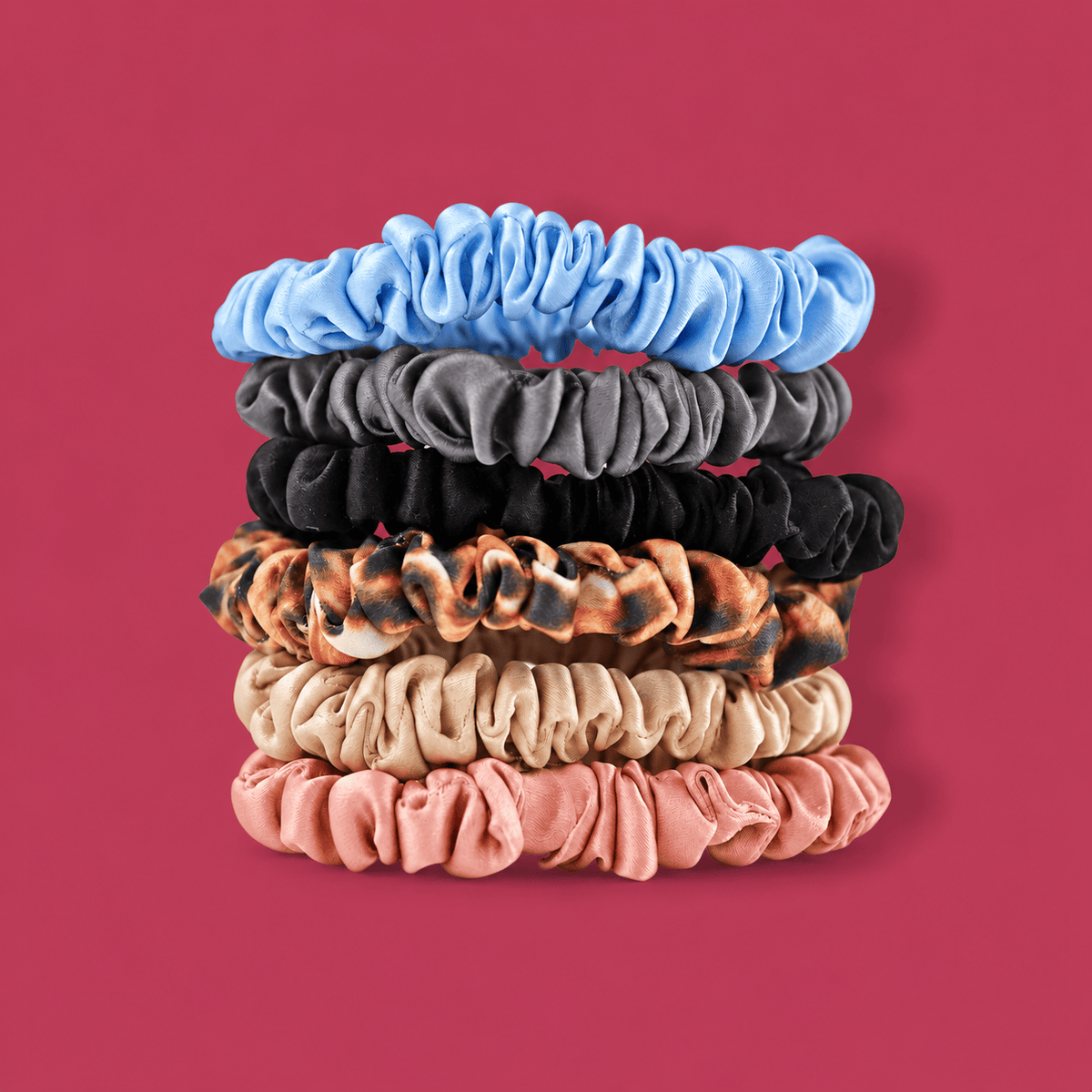 Six Skinny silk scrunchies stacked on a berry background in sky blue, gunmetal, black, leopard, champagne, and blush.