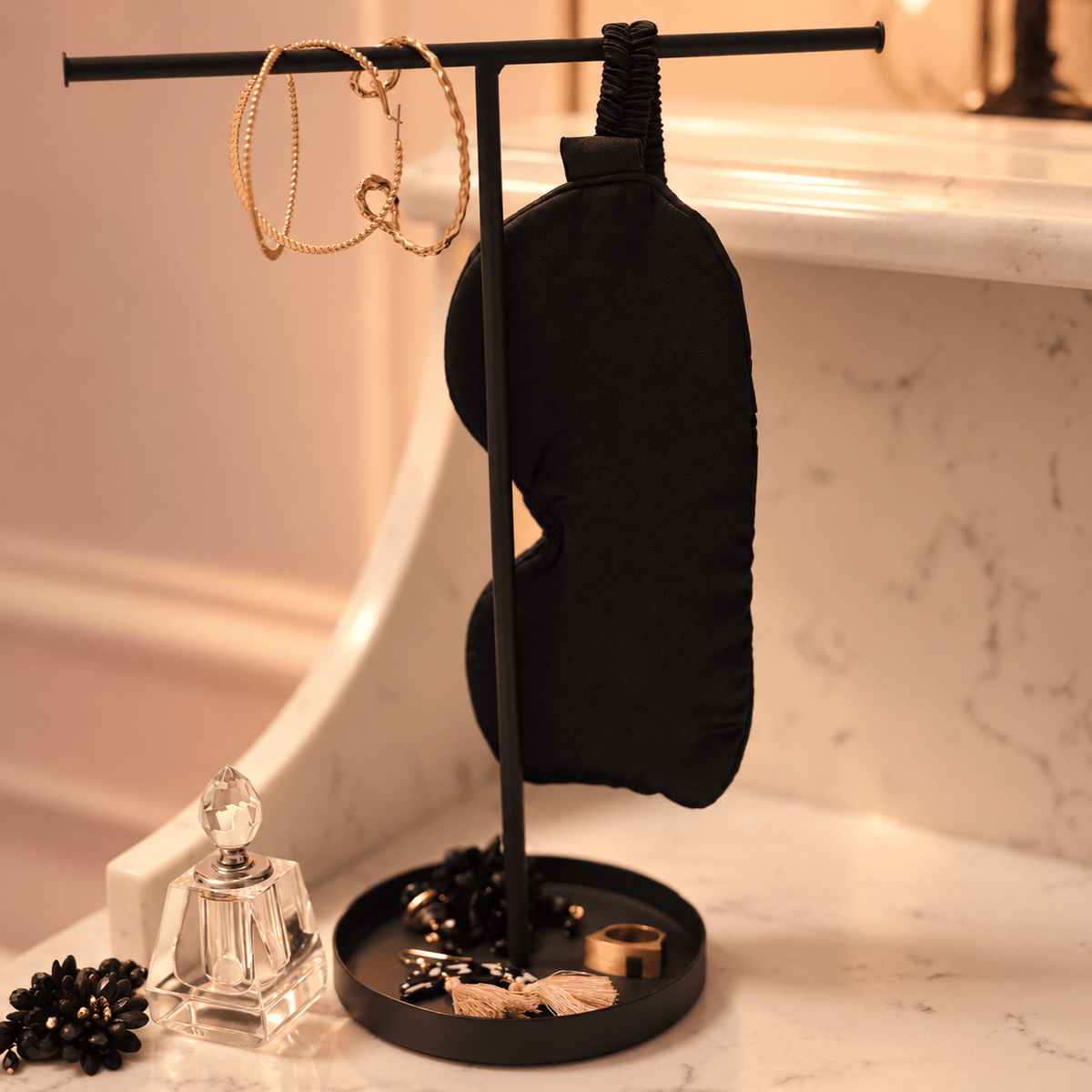 Black satin sleep mask hanging from a jewelry stand on a vanity