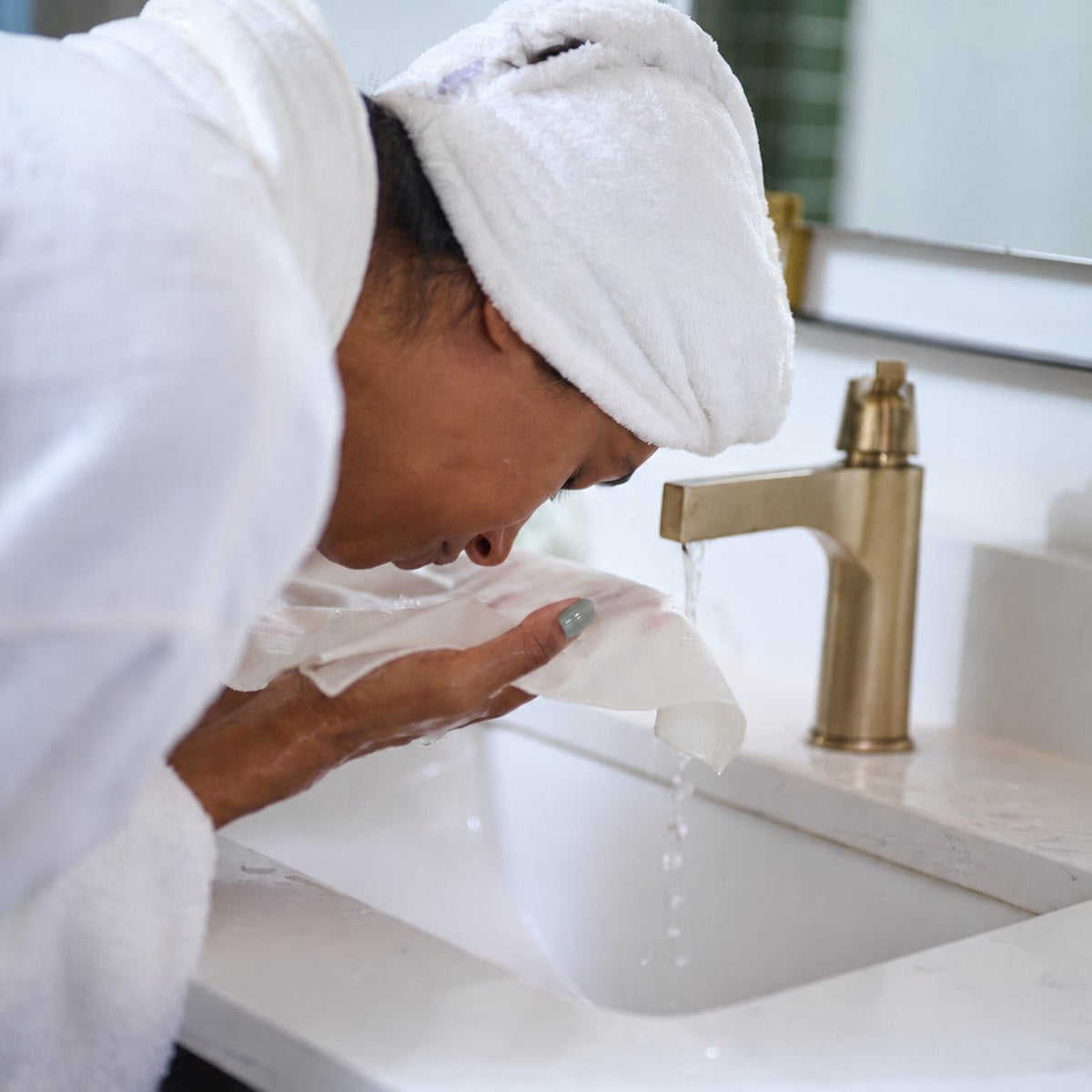 Woman with muted sage nails using the NIGHT raw silk washcloth at a sink.