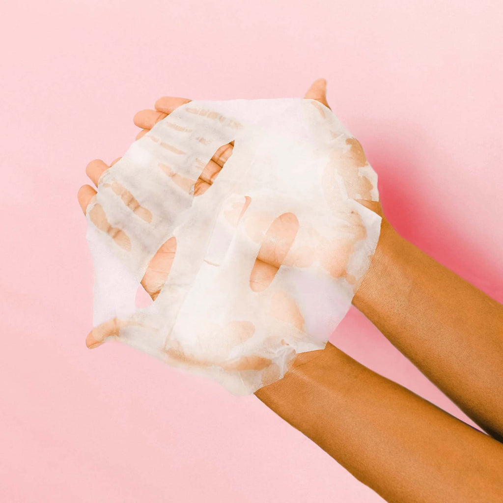 Sheer and wet topical mask being held by hands against a pink background